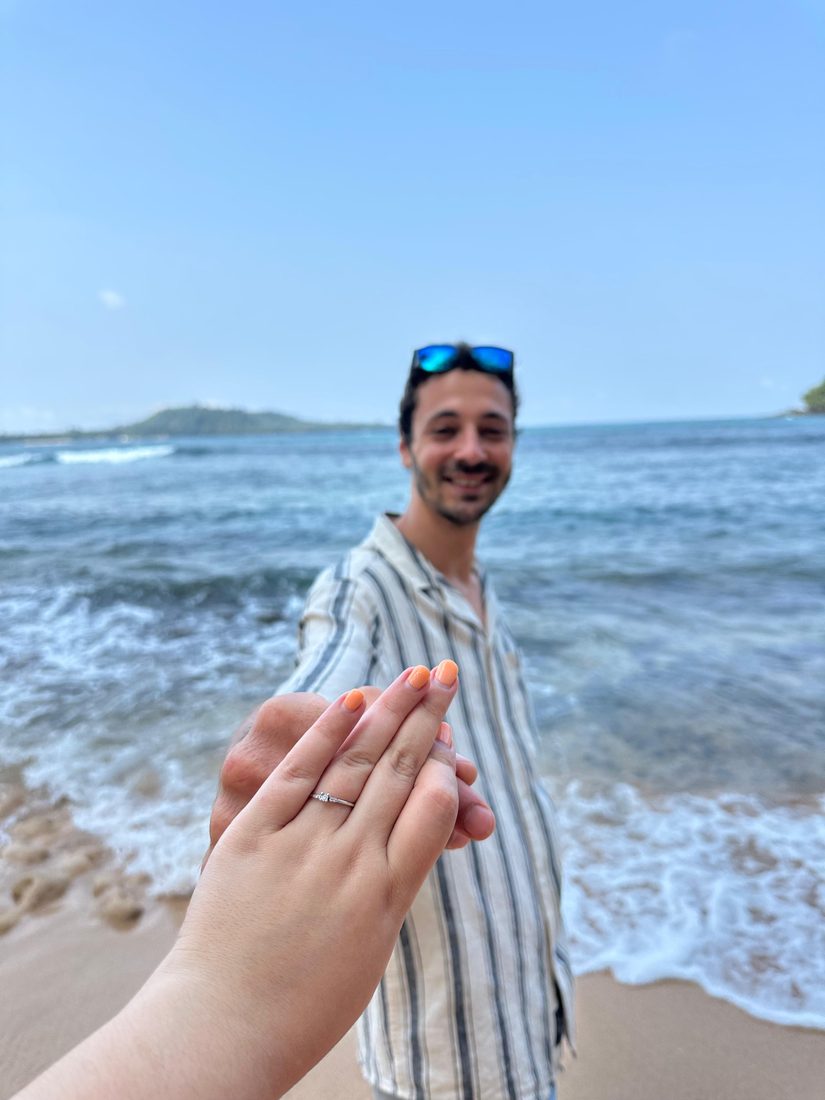 A mão da Inês com o anel, com o Francisco na praia ao fundo